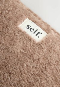 Nærbillede af et beige fleece stof med et hvidt syet label, der viser ordet "self." i sort lille bogstaver.