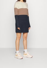 Abito maglione a maglia con strisce orizzontali color crema, marrone chiaro e blu navy, maniche lunghe e vestibilità rilassata. Indossato con sneakers rosa chiaro.