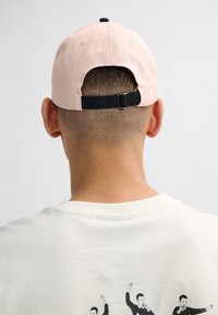 Light pink cotton baseball cap with a black adjustable strap at the back. Visible stitching and a flat peak. White crew neck t-shirt beneath.