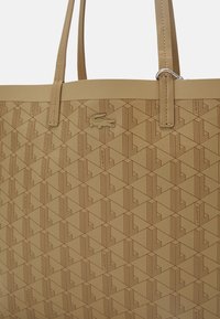 Sac fourre-tout beige en matériau texturé, présentant un motif géométrique et un logo Lacoste embossé. Bandoulières ajustables avec ferrures métalliques.