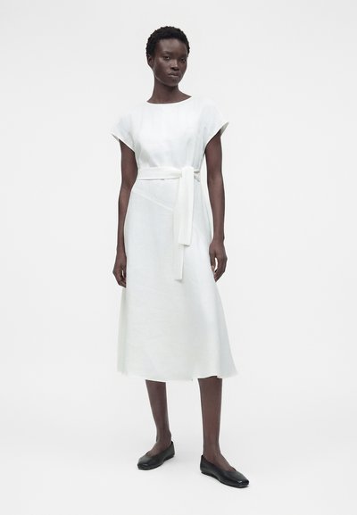 Γυναίκα που στέκεται, φορά ένα άσπρο φόρεμα μέσης με κοντά μανίκια, ζώνη στη μέση και μαύρα επίπεδα παπούτσια, μπροστά σε απλό λευκό φόντο.