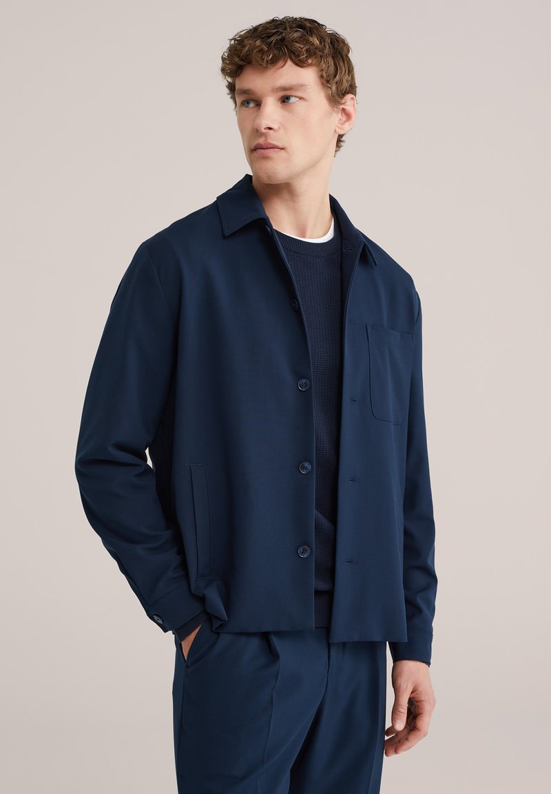 Jaunuolis dėvi tamsiai mėlyną švarkelį su sagomis ant tamsaus megztinio, rankos kišenėse, žiūri į šoną neutralaus fono fone.