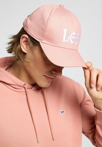 Różowa czapka bejsbolowa z haftowanym białym logo "Lee", składająca się z sześciu paneli i regulowanym paskiem z tyłu. Noszona z dopasowanym hoodie.