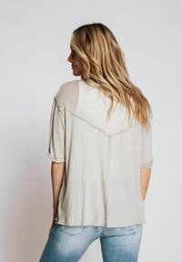 Helle beige, weich strukturierte Bluse mit aufgerollten Ärmeln, die ein geschichtetes Rückendesign und einen fransigen Saum aufweist. Getragen über einer hellblauen Jeans.