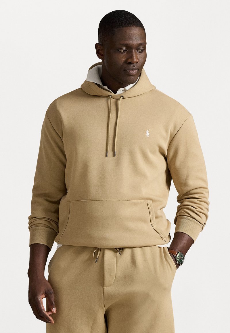 Zandkleurige hoodie met een kangoeroezak, capuchon met trekkoord, geribbelde manchetten en een klein logo op de linkerborst. Soepele, zachte stof. Gecombineerd met bijpassende broek.