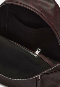 Zaino in pelle marrone scuro con testo del logo rosso e una cerniera parzialmente aperta che mostra una tasca interna nera con un tirante della cerniera argentato.