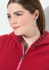 Punainen fleece-huppari, jossa on valkoinen ja punainen ruutukuosi vetoketjun vuorauksessa sekä musta kukka-ompelu kauluksessa. Hopeinen yksityiskohta vetoketjussa.