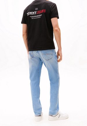 Homme portant un jean bleu clair, des baskets blanches et un t-shirt noir Tommy Jeans avec le logo au dos, debout de dos.