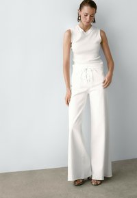 Witte mouwloze top gecombineerd met hoge witte flared broeken met een frontale vetersluiting. Model draagt textuur zwarte hakken.