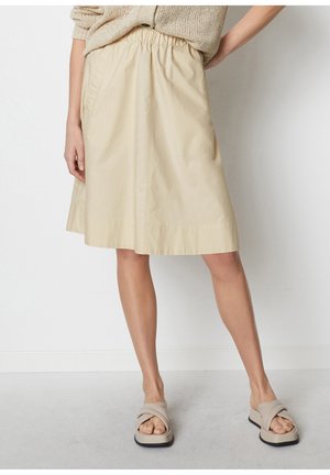 Femme portant une jupe beige mi-genou avec une ceinture élastique, un pull en tricot et des sandales beige à brides croisées, debout contre un mur blanc.