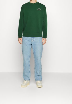 Man draagt een donkergroene trui met krokodillogo, lichtblauwe spijkerbroek en witte sneakers, staand tegen een effen witte achtergrond.