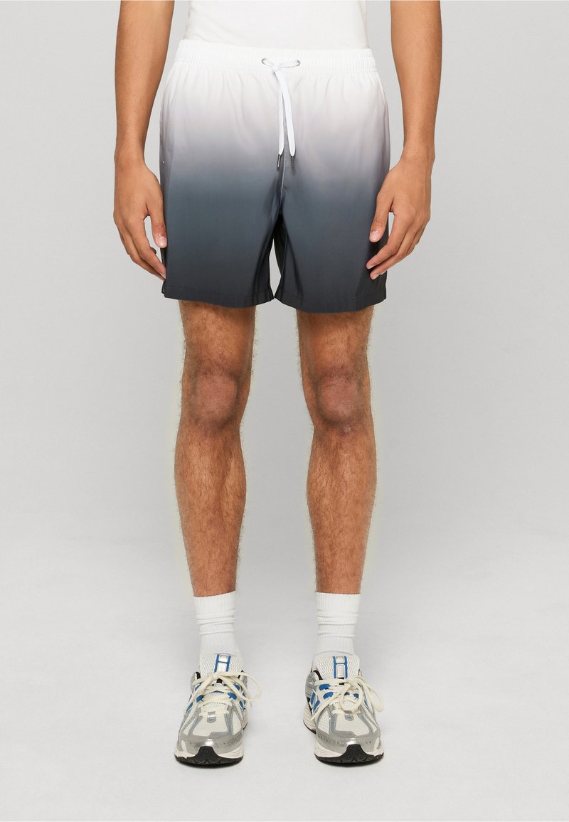 Homme portant un short dégradé de blanc à gris foncé, des chaussettes blanches et des chaussures de sport grises, debout devant un fond gris uni.