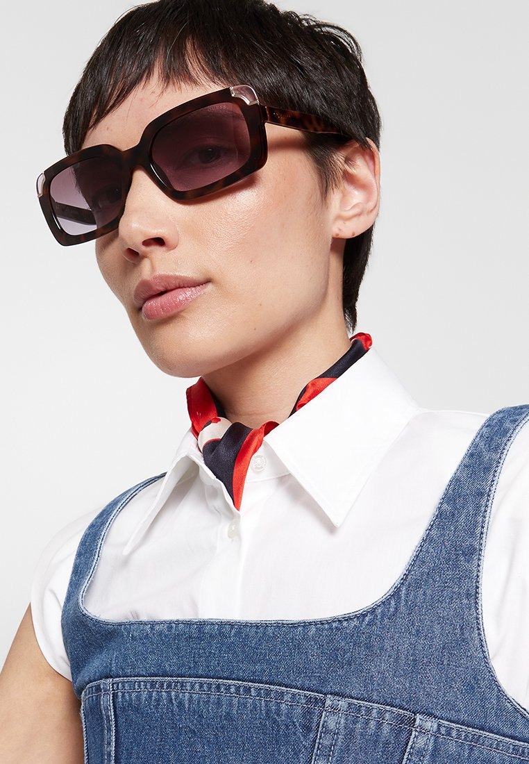 Azetat-Sonnenbrille mit großen rechteckigen Rahmen in dunklem Schildpatt und Verlaufsgläsern. Das Model trägt ein weißes Hemd und Jeansoveralls.