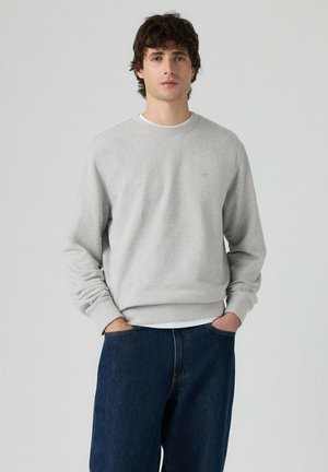 Jeune homme aux cheveux bouclés portant un sweat-shirt gris clair à manches longues et un jean bleu foncé, les mains dans les poches, debout devant un fond uni.