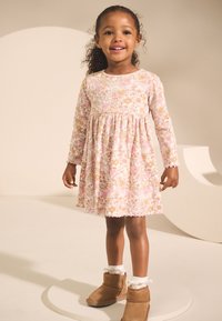 Rosa blommig klänning med långa ärmar och veckad kjol, med vågig nederkant, bärs med bruna ankelboots och vita volangstrumpor.