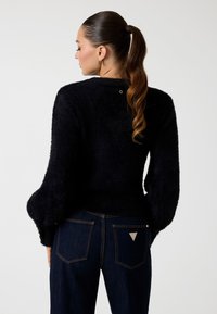 Suéter negro de felpa con mangas voluminosas, dobladillo ajustado y un detalle del logo dorado en la parte posterior. Combinado con jeans azul oscuro que tienen acentos de logo.