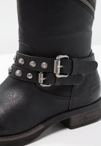 Schwarzer Leder-Stiefelette mit strukturiertem Obermaterial, einem dekorativen Riemen mit silbernen Nieten und einem festen Blockabsatz. Reißverschlussdetail an der Seite.