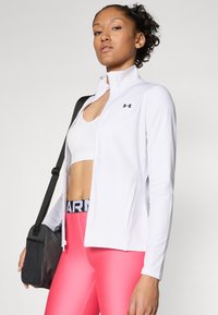 White athletic jacket with a zip front and high collar, paired with pink leggings. Black logo on the jacket, bag slung over the shoulder.