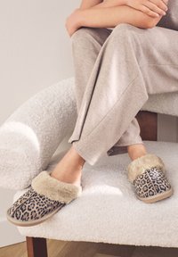 Chaussons à imprimé léopard avec doublure en fausse fourrure douce et semelle beige, portés avec un pantalon de détente gris clair sur un fauteuil blanc texturé.