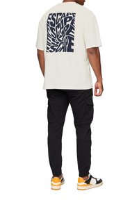 Witte t-shirt met korte mouwen en een grote marineblauwe grafische print met de tekst "ESCAPE" op de rug. Gecombineerd met zwarte cargobroeken en multikleurige sneakers.