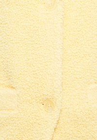 Veste boutonnée crème jaune avec un tissu texturé et duveteux. Dotée d'une fermeture à boutons et de deux poches avant.