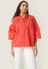 Camicia oversize corallo realizzata in cotone, con maniche a sbuffo, bottoni frontali e dettagli arricciati attorno al collo. Abbinata a pantaloni bianchi.