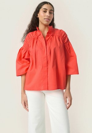 Camisa de gran tamaño en coral, hecha de algodón, con mangas abullonadas, botones frontales y detalles fruncidos alrededor del escote. Combinada con pantalones blancos.