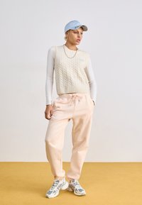 Top sin mangas de punto trenzado en color crema, pantalones deportivos rosa claro con cordón, gorra azul y zapatillas multicolores con acentos. Fondo liso.