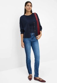 Marineblaue Rippstrickjacke mit braunen Knöpfen, kombiniert mit einem marineblauen Oberteil und blauen Jeans mit Seitenstreifen. Getragen mit burgunderfarbenen Ballerinas.