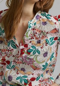 Blouse florale en tissu léger. Présente des motifs vibrants rouges, roses et verts avec des fleurs et des feuilles détaillées. Design à col boutonné.