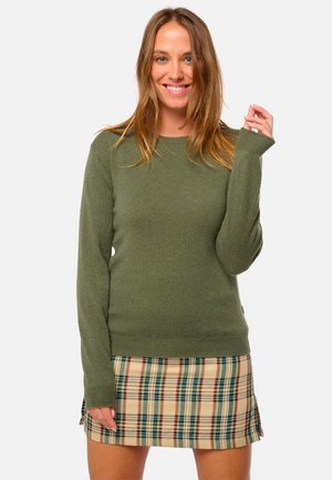 Pull en tricot vert olive avec de longues manches et des poignets côtelés, associé à une jupe écossaise beige courte avec des lignes vertes, rouges et jaunes.