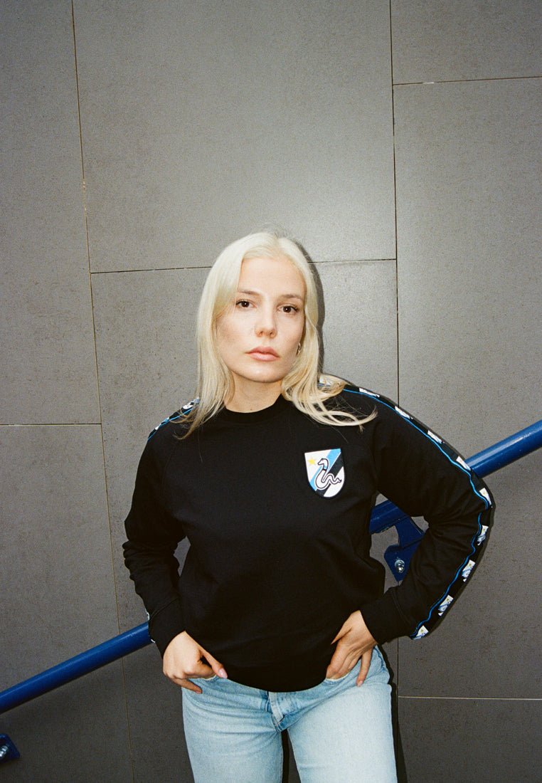 Zwarte sweatshirt met lange mouwen, voorzien van een blauw-wit schildlogo en blauwe strepen langs de mouwen, gecombineerd met lichtblauwe jeans.