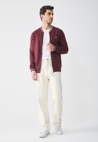 Bordo zip-up jakna s rebrastim ovratnikom i manžetama, uparena s bijelom majicom i krem hlačama. Model nosi bijele tenisice.