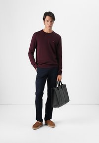 Pulover burgund, pantaloni bleumarin și pantofi slip-on maro. Geantă de piele neagră cu design structurat. Modelul stă în picioare pe un fundal alb.