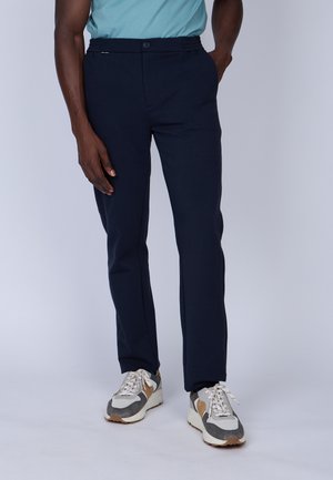 Pantalon bleu marine avec une texture douce, coupe slim, fermeture à bouton et taille élastique. Associé à des baskets de sport grises et beiges.
