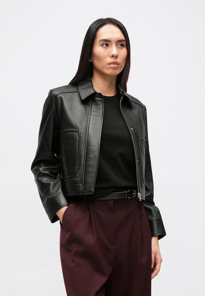 Čierna kožená bunda s golierom, predným zipsom a bočnými vreckami, nosená cez čierny sveter a vínové nohavice. Hladký, textúrovaný povrch.