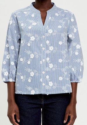 Femme portant un chemisier à rayures bleu et blanc avec une broderie florale blanche et des manches trois-quarts, associé à un jean foncé.