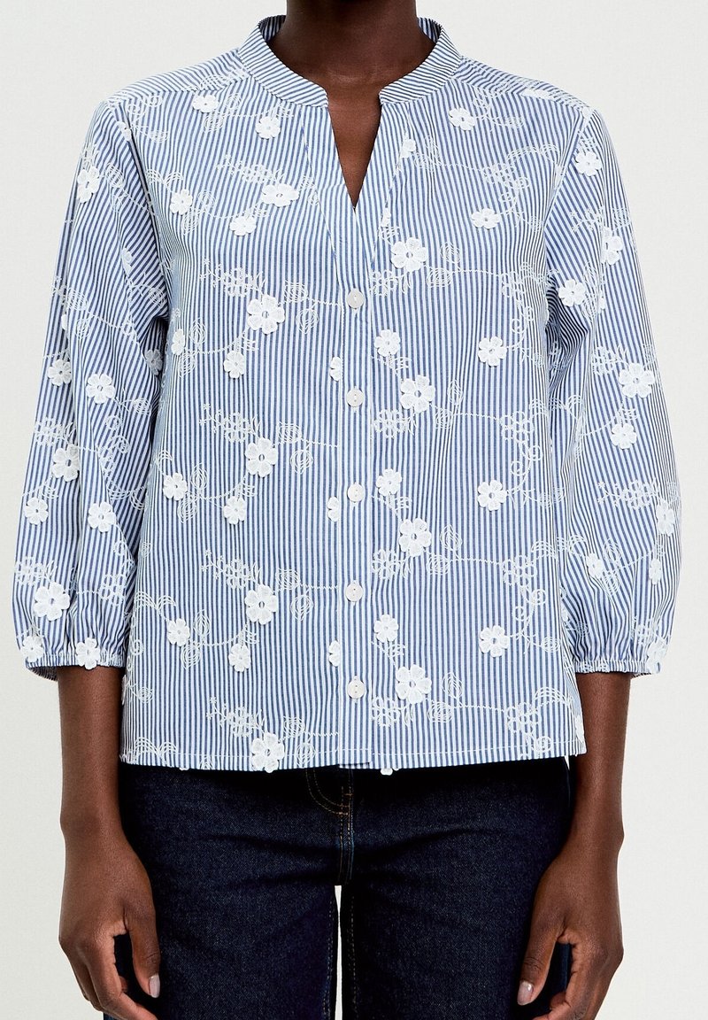 Femme portant un chemisier à rayures bleu et blanc avec une broderie florale blanche et des manches trois-quarts, associé à un jean foncé.
