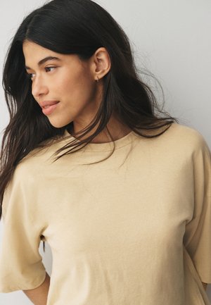 Ung kvinde med langt mørkt hår iført en beige oversized skjorte, kigger let til venstre med et roligt udtryk mod en ensfarvet baggrund.