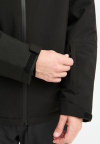 Zwarte waterdichte jas met een aangesloten mouw en verstelbare klittenband manchetten, en een ritszak aan de zijkant. Gladde textuur en gestroomlijnd ontwerp.