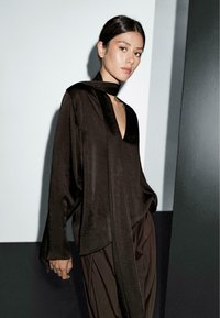 Blouse noire en soie avec un décolleté en V et des manches longues, présentant un design drapé et un détail de foulard au cou. Texture sans couture et coupe fluide.