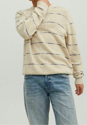 Person iført en beige sweater med tynde mørke horisontale striber og lyseblå slidte jeans, hånd hvilende på nakken.
