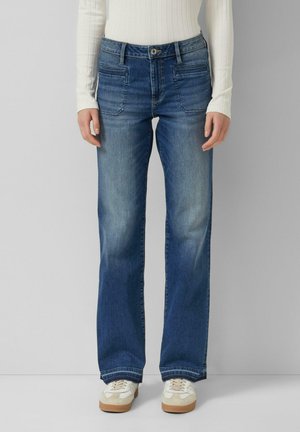 Osoba nosząca proste, niebieskie dżinsy o średnim odcieniu z przednimi kieszeniami naszywanymi oraz białe trampki, stojąca na tle jednolitego jasnego tła.