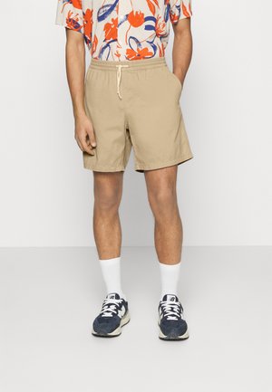 Beige shorts with an elastic waistband and drawstring, featuring two side pockets. Worn with white crew socks and navy sneakers.