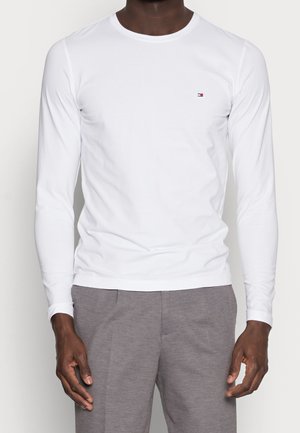 Langärmeliges weißes T-Shirt aus Baumwolle mit rundem Ausschnitt und kleinem Logo oben links. Glatte Textur, figurbetonte Passform.