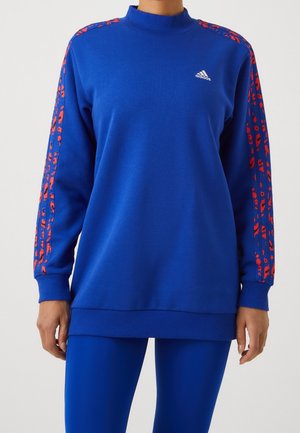 Kobieta ubrana w niebieską bluzę Adidas z czerwonymi paskami w panterkę na rękawach oraz dopasowane niebieskie legginsy na białym tle.