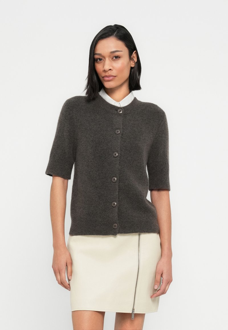 Σκούρο καφέ κοντομάνικο ζακετάκι από μαλακό υλικό με στρογγυλή λαιμόκοψη και επτά κουμπιά, φορεμένο πάνω από ένα λευκό πουκάμισο και ανοιχτόχρωμη φούστα.