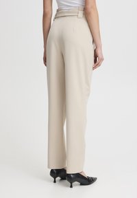 Pantaloni beige a vita alta con un design sartoriale, dotati di una cintura e gamba dritta, abbinati a tacchi neri a punta.