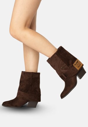 Benen met donkerbruine suède laarzen tot halverwege de kuit met lage gestapelde hakken en omgeslagen boorden, één voet opgetild alsof er een stap naar voren wordt gezet.