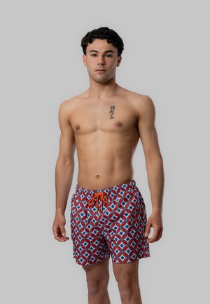 Giovane uomo in piedi senza maglietta che indossa pantaloncini da bagno rossi, bianchi e blu a fantasia con cordoncino arancione su sfondo grigio semplice.